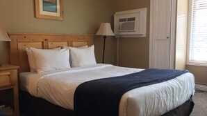 A neatly made bed with white bedding and a dark blue throw, a wooden headboard, a framed picture on the wall, a standing lamp, and a window with blinds. - Cute cozy condo near ski resorts (Park City)