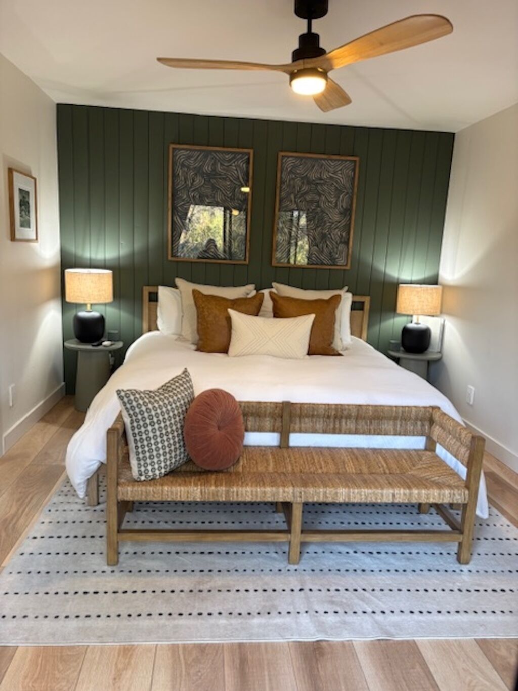 A bedroom with a wooden bench, a bed with multiple pillows, two wall-mounted lamps, and a ceiling fan.