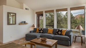 Un salon moderne avec un canapé d’angle gris, une table basse en bois et une table à manger. De grandes fenêtres offrent une vue sur les arbres et les montagnes.