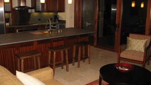 A modern kitchen with a bar area, wooden bar stools, a dining chair, and a small table with a bowl.