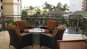 A balcony with wicker furniture, a glass table, and a view of palm trees and buildings.