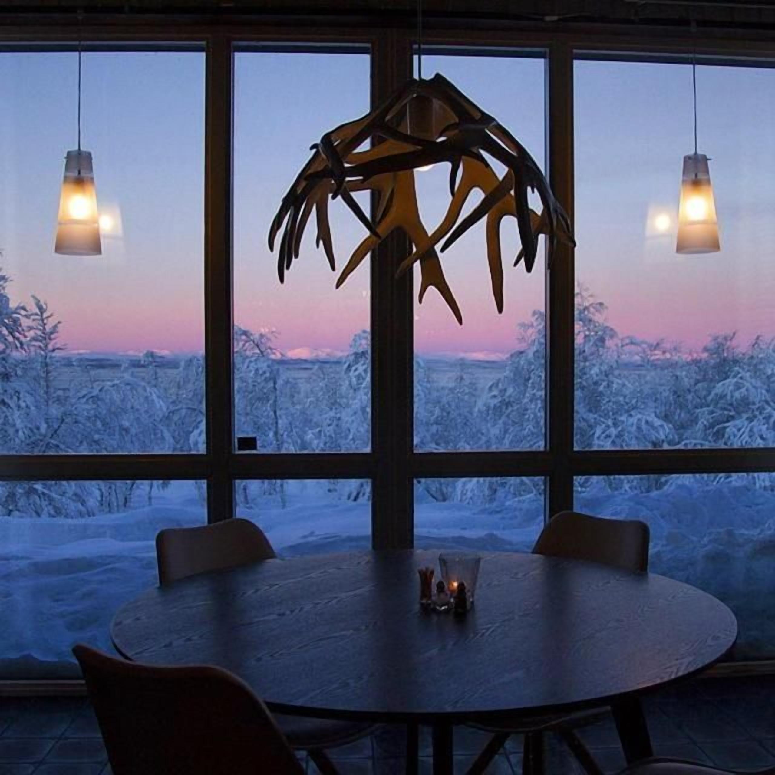A room with a round table and chairs, a hanging light fixture, and a view of a snowy landscape through large windows.