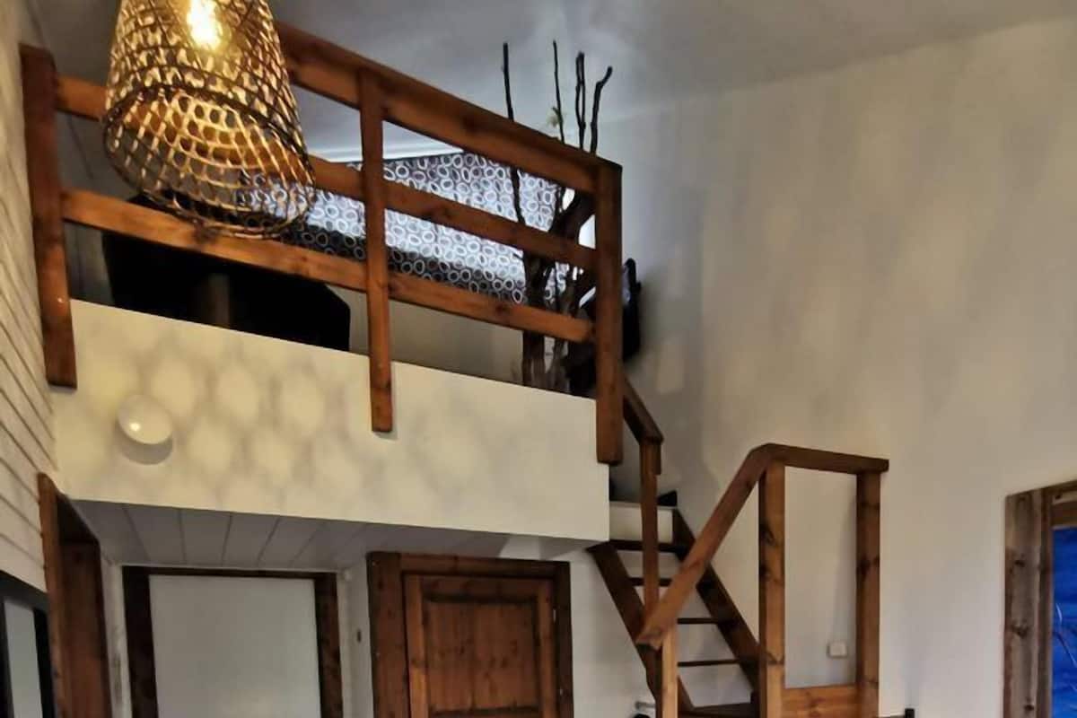 Ett sovrum på entresolplan med en trätrappa, en säng med ett vitt överkast och en hängande lampa.