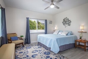 Une chambre à coucher comprenant un lit, une chaise, une petite table, un ventilateur de plafond, une fenêtre avec des rideaux et un luminaire fixé au mur.