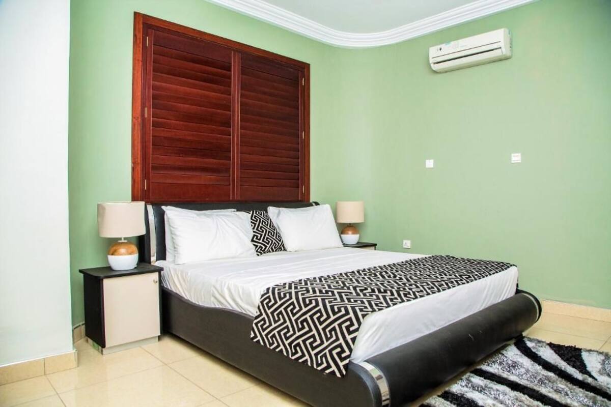 A bedroom with a bed, two bedside tables, a nightstand, a patterned throw blanket, a wooden headboard, and an air conditioning unit.