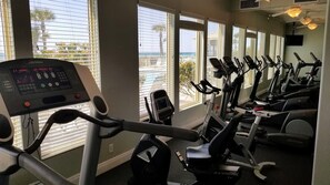 Un gimnasio con varias bicicletas estáticas y una máquina de cardio, ventanas amplias con persianas y vistas al océano.