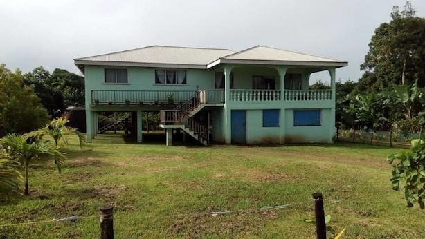 A two-story house with a balcony, surrounded by a grassy yard and trees. - Lady Maria's Place (Pangai)
