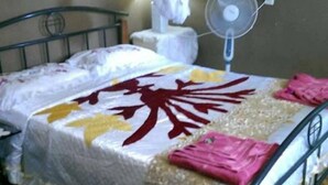 A single bed with a white bedspread and a decorative red pattern, a bedside fan, and a mosquito net above the bed. - Lady Maria's Place (Pangai)