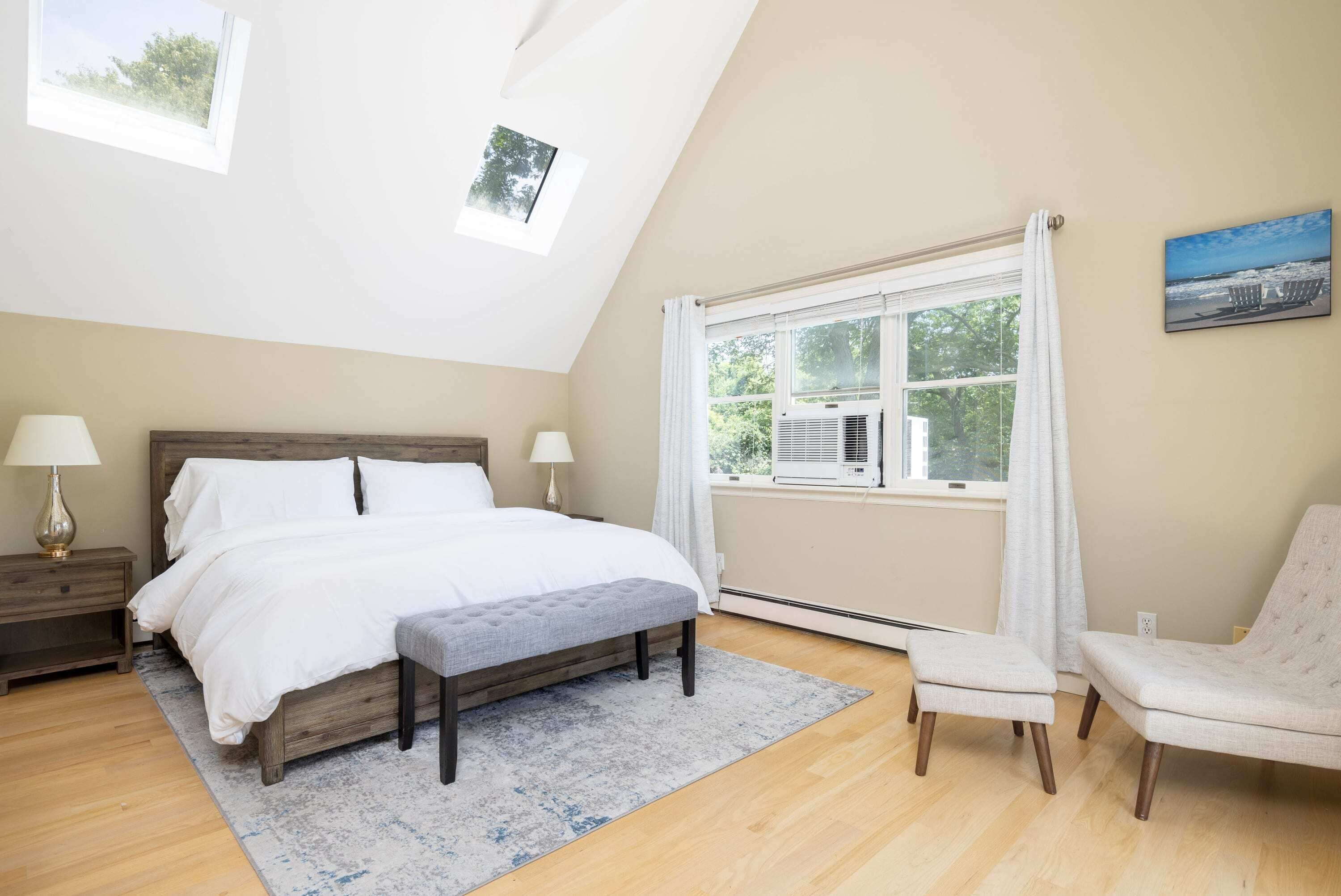A bedroom with a large bed, a bench, a chair, a nightstand, and a window with curtains.