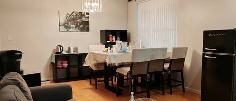 A modern living room with a sofa, a dining table with chairs, a coffee machine, a refrigerator, and a chandelier.