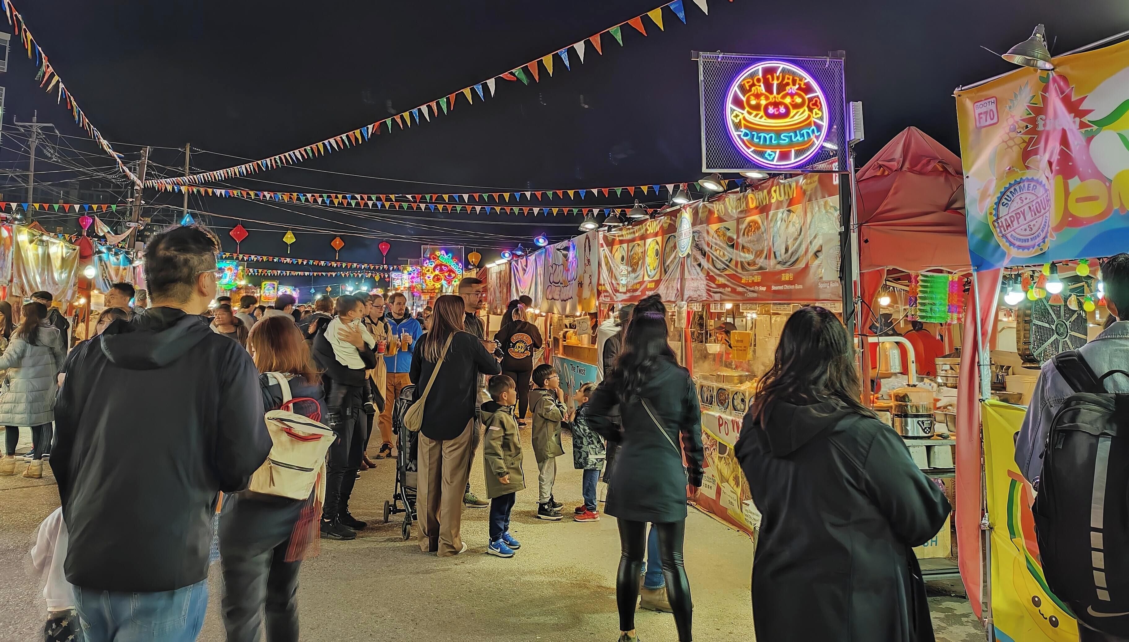 A crowded outdoor market with various stalls and a neon sign.