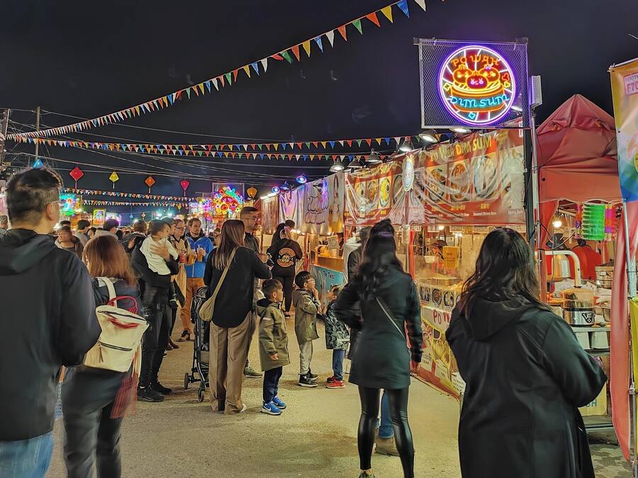 A crowded outdoor market with various stalls and a neon sign.
