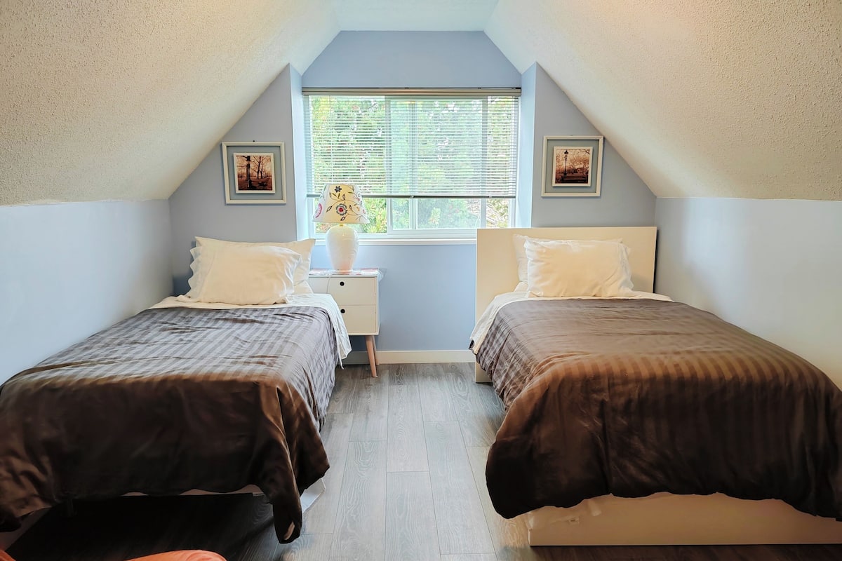 A bedroom with two beds, a window with blinds, a nightstand with a lamp, and framed pictures on the walls.