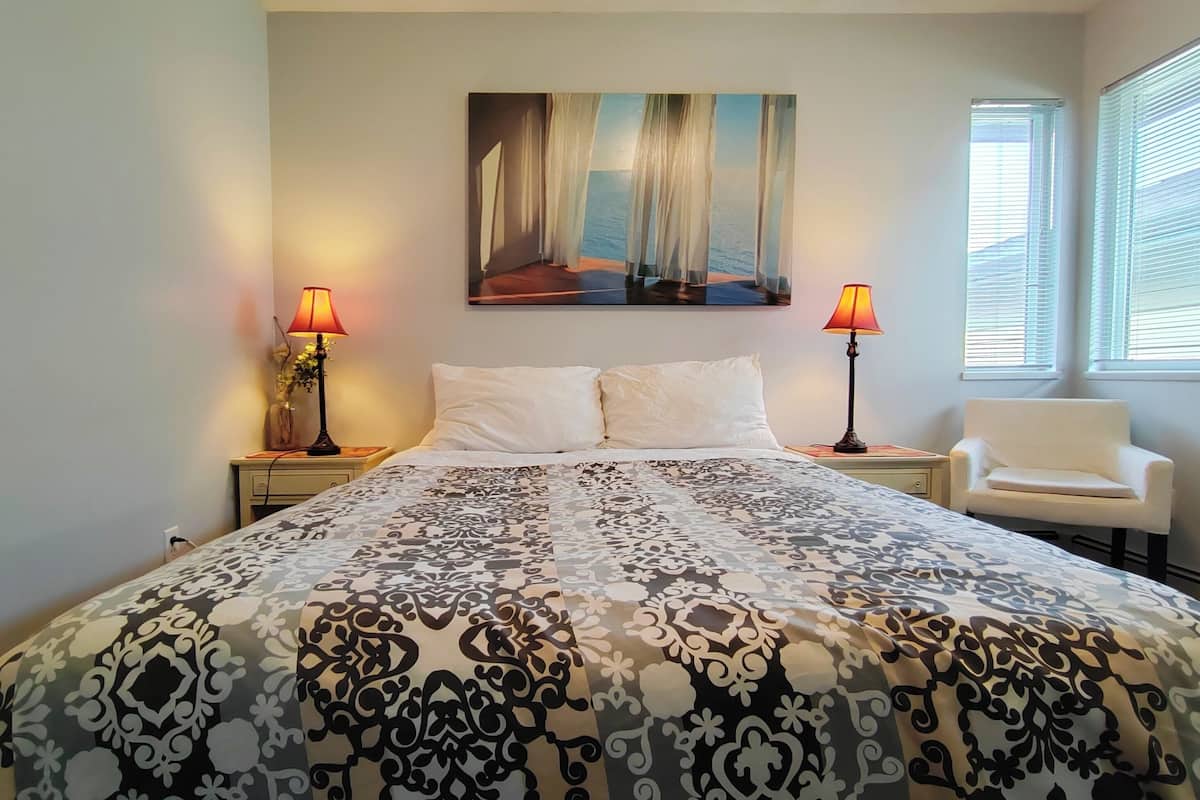 A bedroom with a bed, two bedside lamps, a chair, a painting on the wall, and a window with blinds.