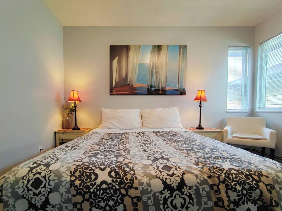 A bedroom with a bed, two bedside lamps, a chair, a painting on the wall, and a window with blinds.