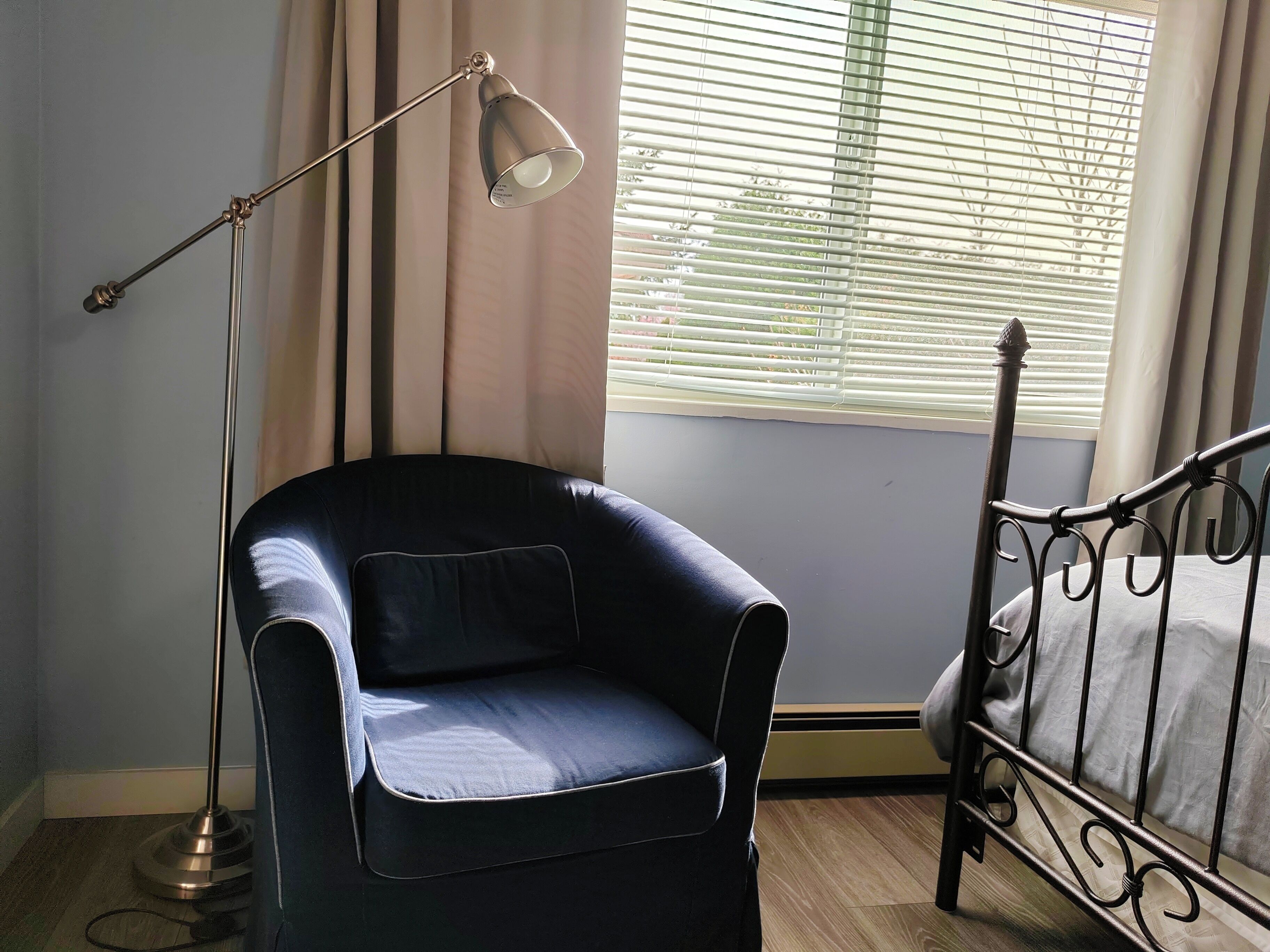 A room with a single black chair, a standing floor lamp, a bed with metal headboard, and a window with blinds.
