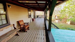 Una terraza de madera con banco y mesa, un área cubierta con barandilla, y vista a un jardín y una casa al fondo.