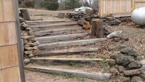 Una escalera de madera que sube hacia una casa, rodeada por un muro de piedra y una cerca de madera.