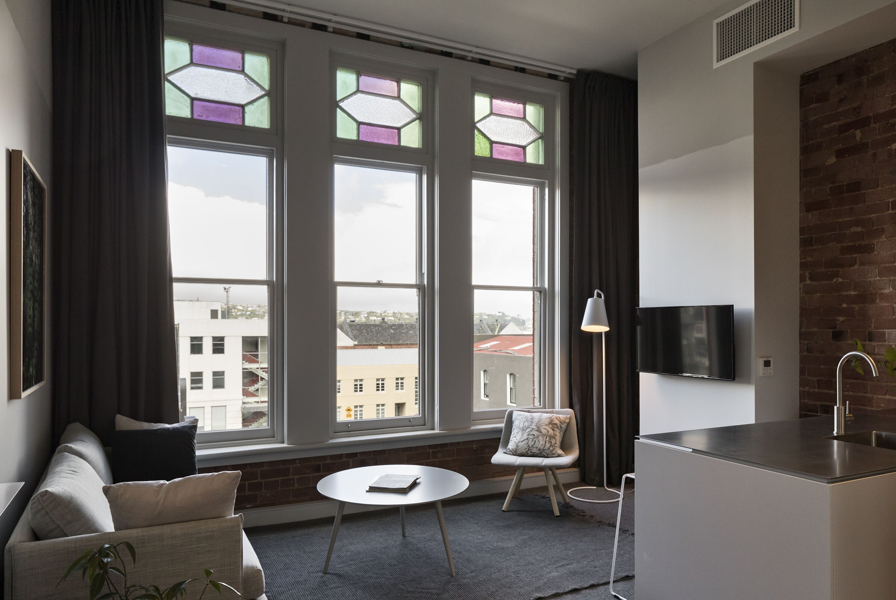 A modern living room with a sofa, a small round table, a chair, a kitchen area, and large windows with stained glass.
