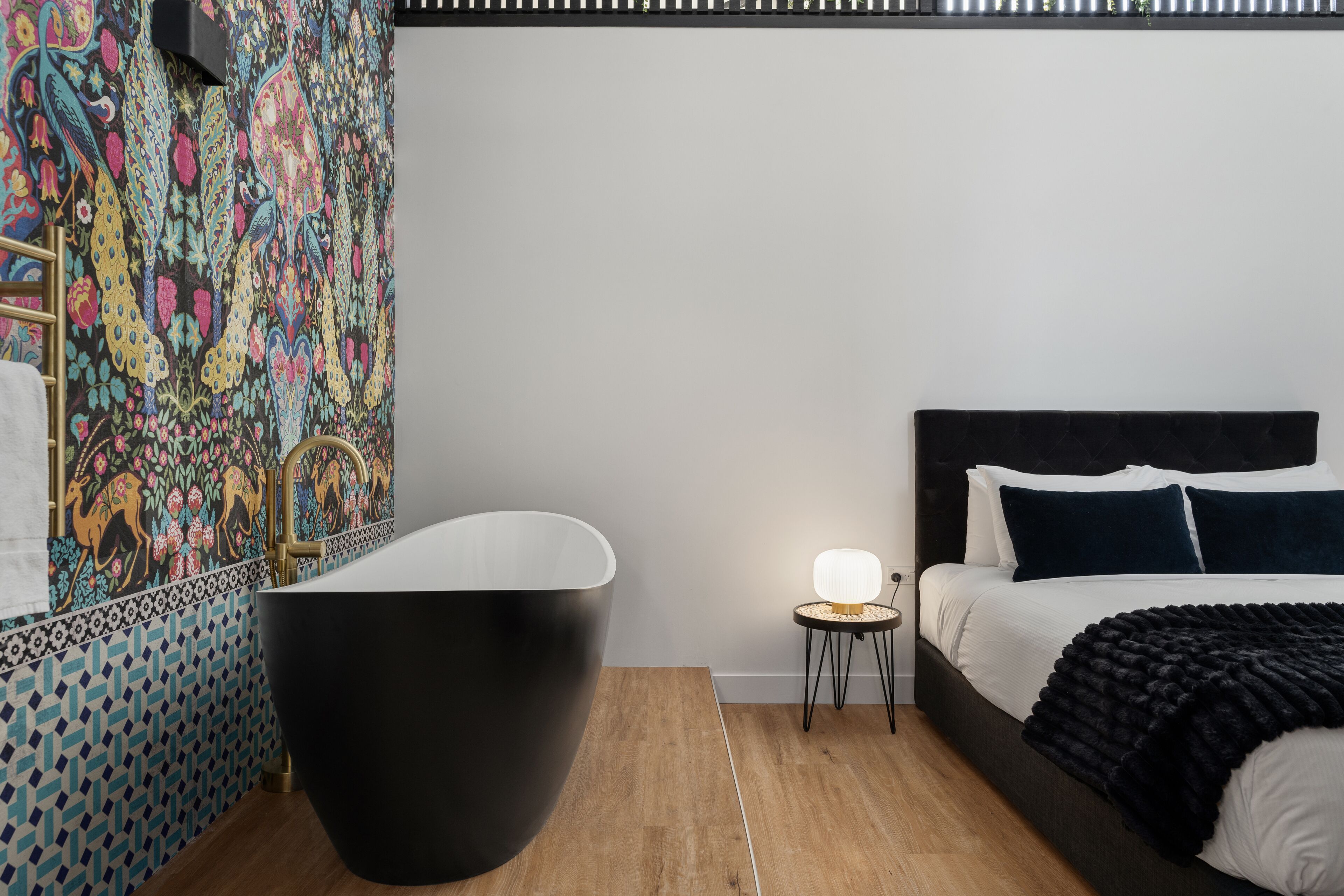 A modern bathroom with a freestanding bathtub, a decorative wall with a floral pattern, and a bedside table with a lamp.