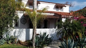 Un bâtiment blanc avec un toit en tuiles, entouré de plantes tropicales et bordé d’un chemin de pierre.