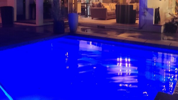 Vue sur une maison depuis la piscine, avec un espace de vie ouvert, un coin repas et une cuisine extérieure visible.