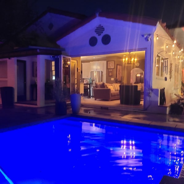 A poolside view of a house with an open living area, a dining area, and a visible outdoor kitchen.