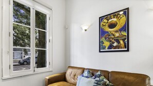 A room with a brown leather sofa, a window with a view of the street, and a framed picture on the wall.