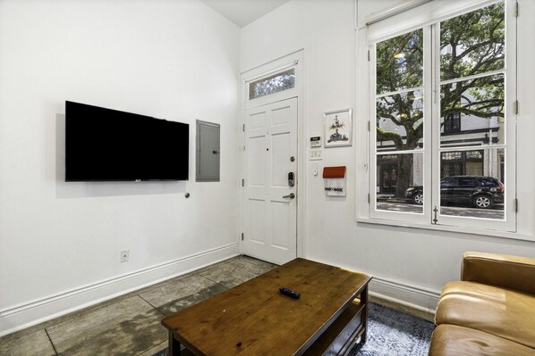 A room with a flat-screen TV mounted on the wall, a wooden coffee table, a leather sofa, a door with a window, and an intercom system.