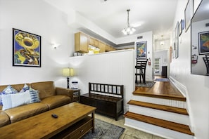 A modern living room with a brown leather sofa, a wooden coffee table, and a small side table with a lamp. There is a staircase leading to an upper level with wooden flooring.