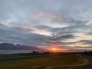 Auringonlasku vuoristomaiseman yllä, ja maisemassa näkyy tie, joka johtaa rakennukseen.