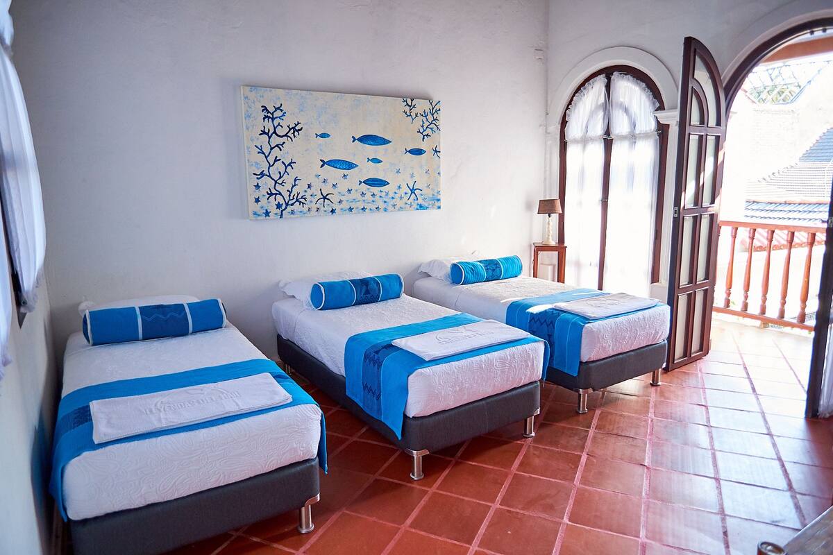 A room with two beds, each with blue and white bedding, a painting of fish and coral on the wall, and a balcony with a railing.
