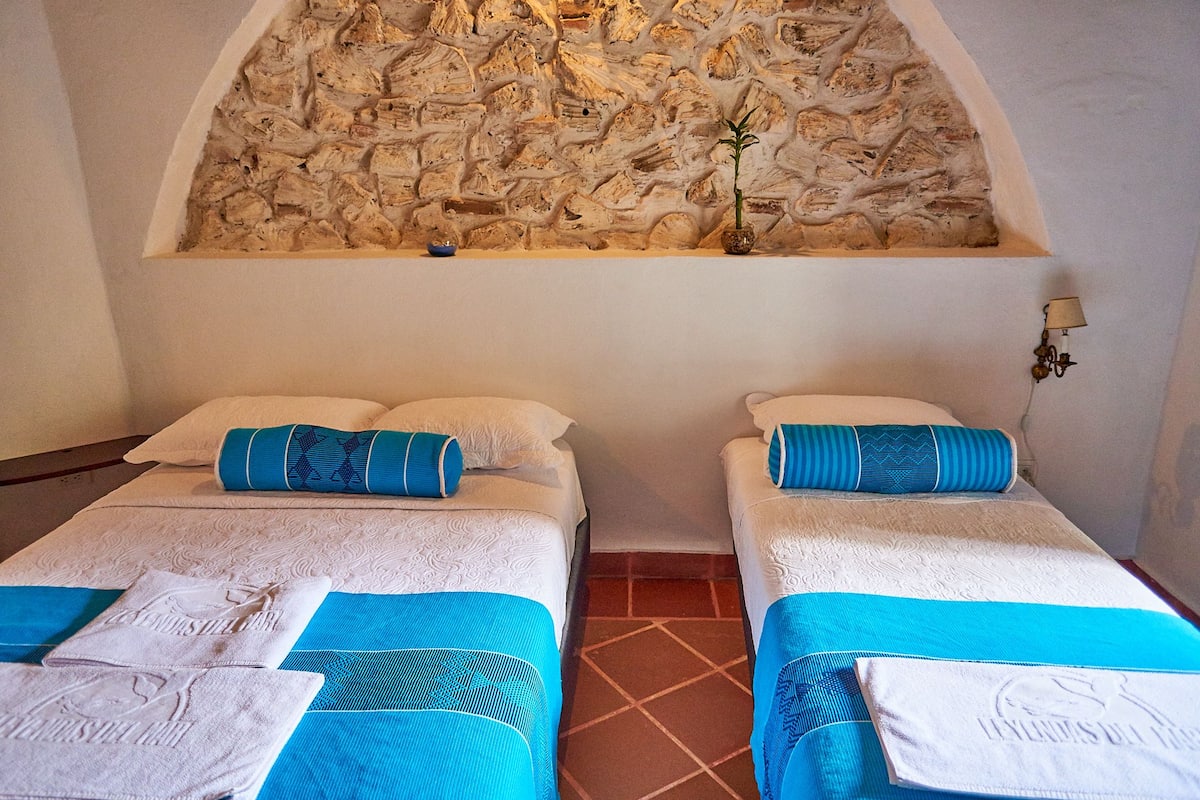 A room with two beds, each with a blue and white patterned bedspread, a small plant on the wall, and a tiled floor.