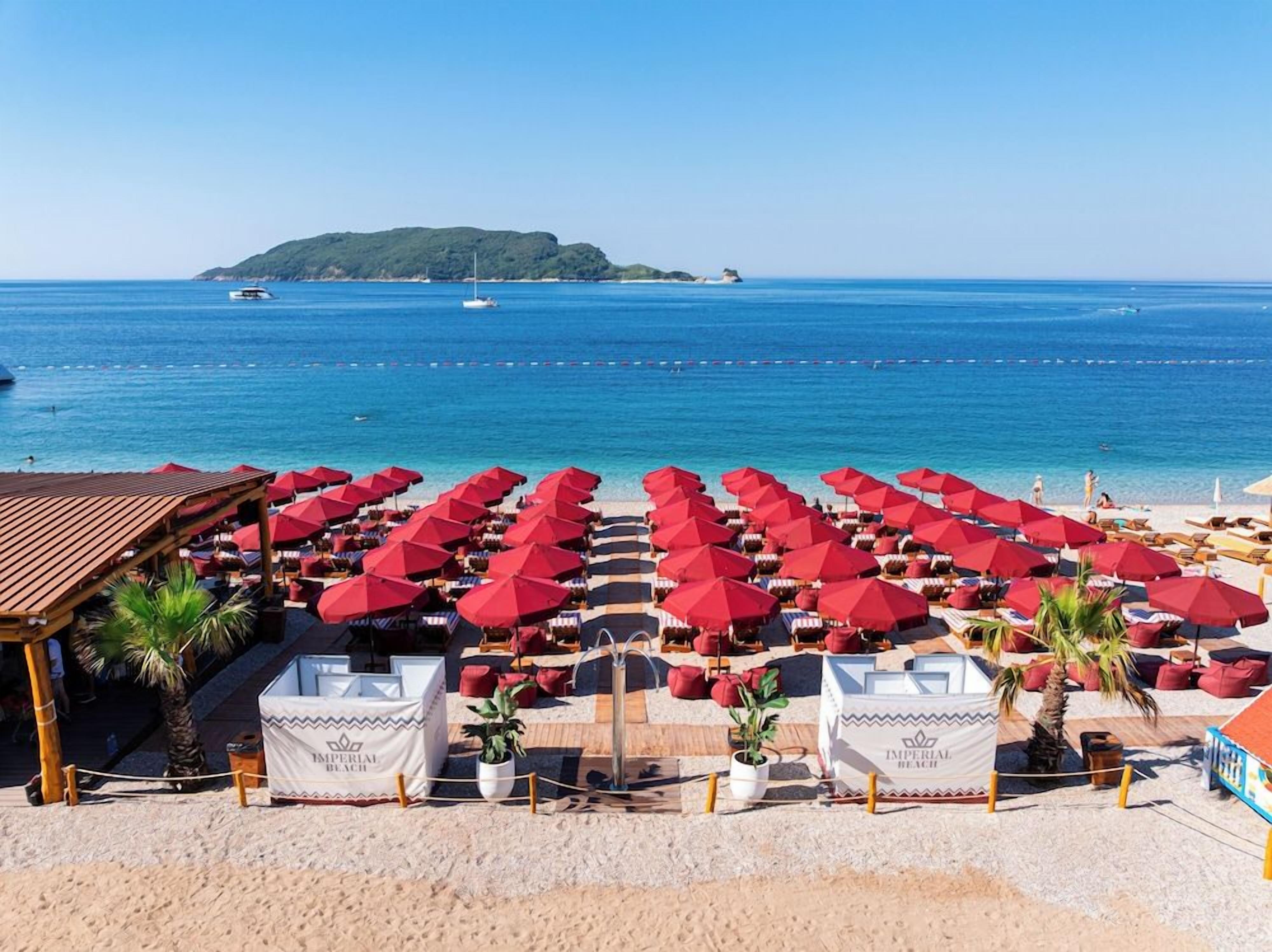 A beach with rows of red umbrellas and lounge chairs, a wooden structure, and a clear view of the sea and a distant island.