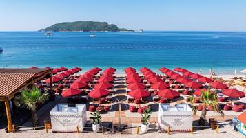 A beach with rows of red umbrellas and lounge chairs, a wooden structure, and a clear view of the sea and a distant island.