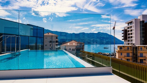 Une piscine sur le toit, entourée d’une paroi en verre, offrant une vue sur une ville côtière et les montagnes.