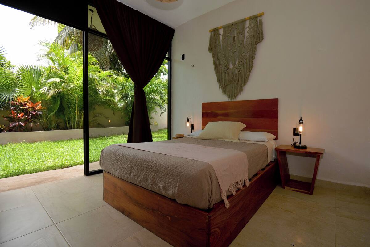 Un dormitorio con una cama grande, una mesita de noche, un adorno macramé en la pared y vistas al exterior.