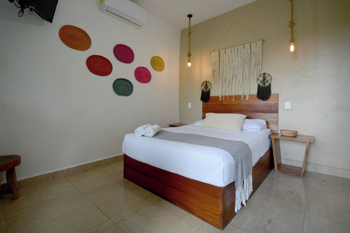 Una cama bien hecha con ropa blanca y un cabecero de madera, una mesita de noche con un cuenco, y una pared decorada con arte circular.