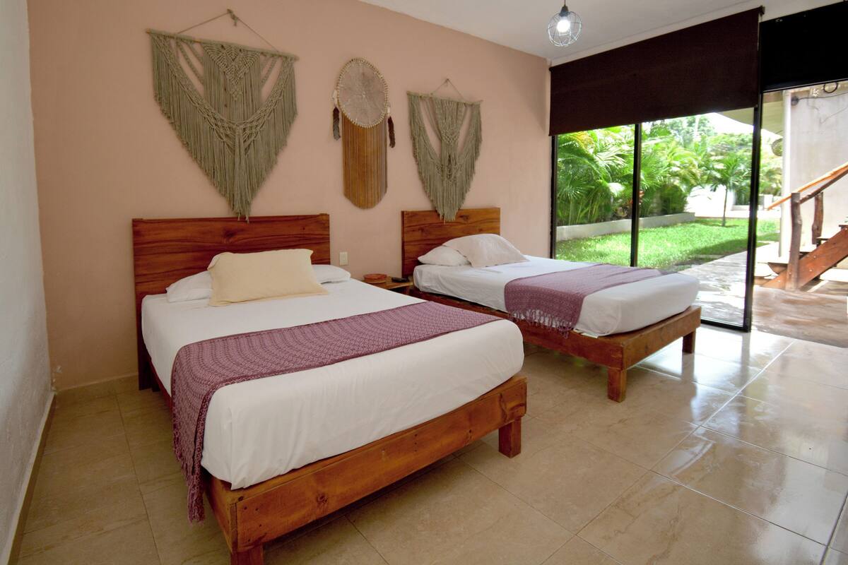 Habitación de hotel con dos camas de madera, un ventanal con vistas a la vegetación y macramés decorativos en la pared.