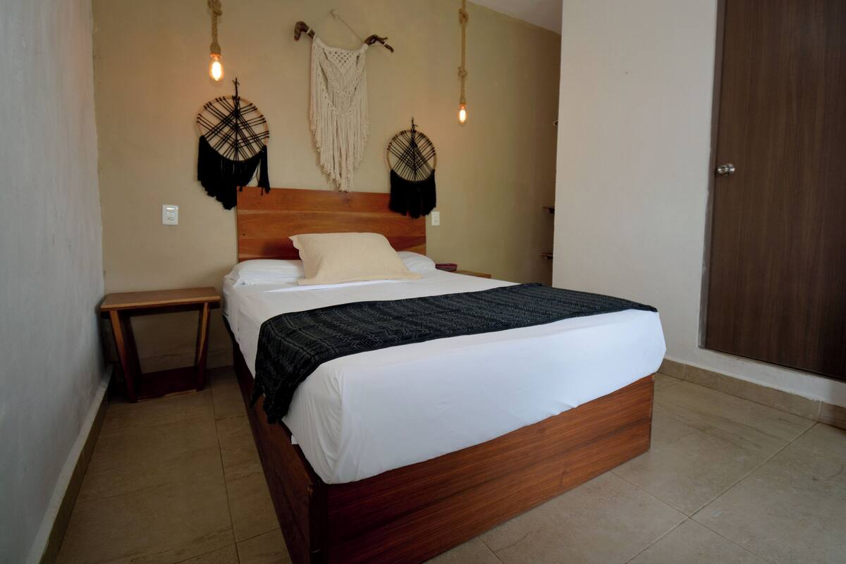 Una cama bien tendida con cabecera de madera, una mesita de noche y dos tapices de macramé colgados en la pared.