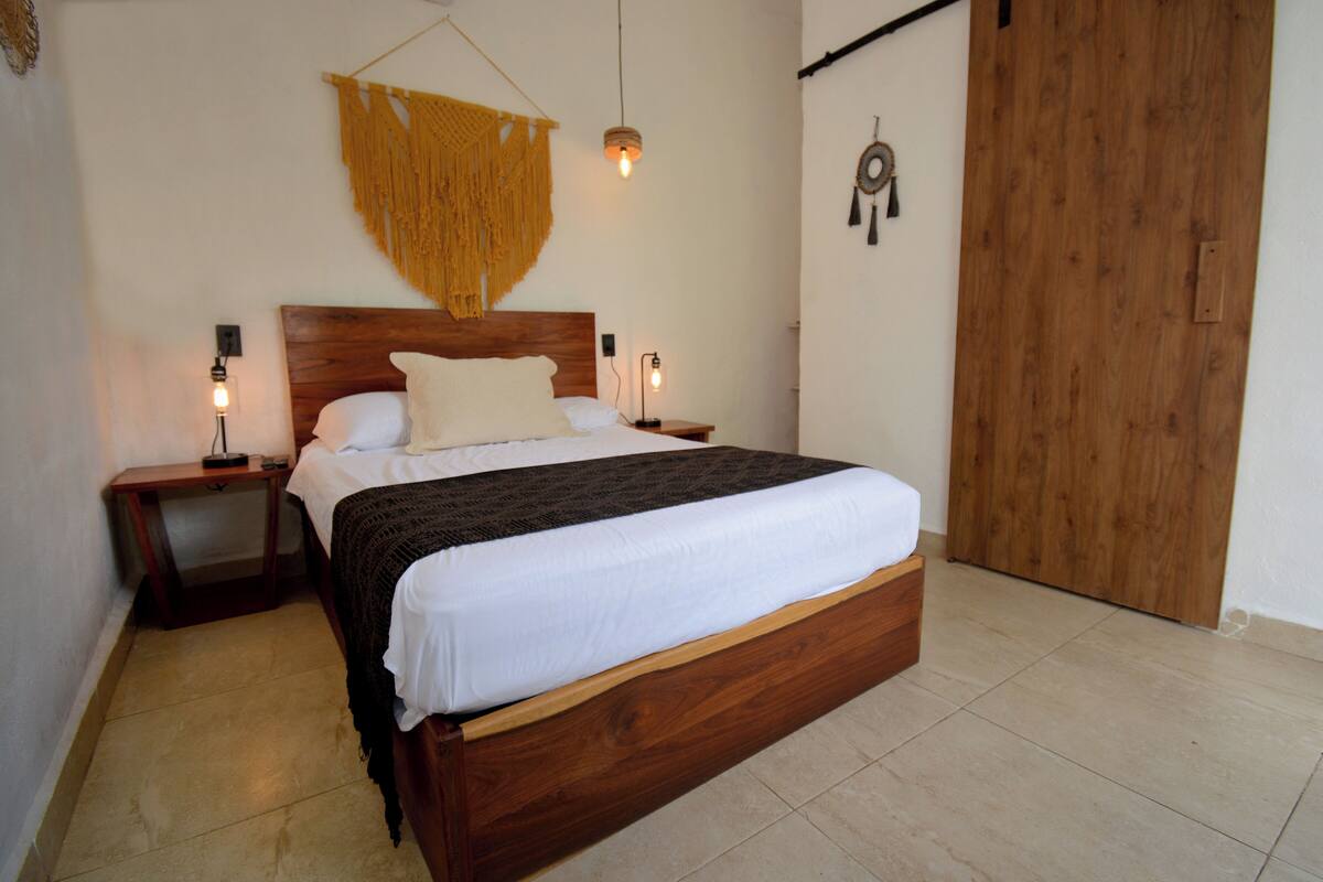 Una cama bien hecha con cabecera de madera, dos mesitas de noche con lámparas, un colgador de macramé y un atrapasueños en la pared.