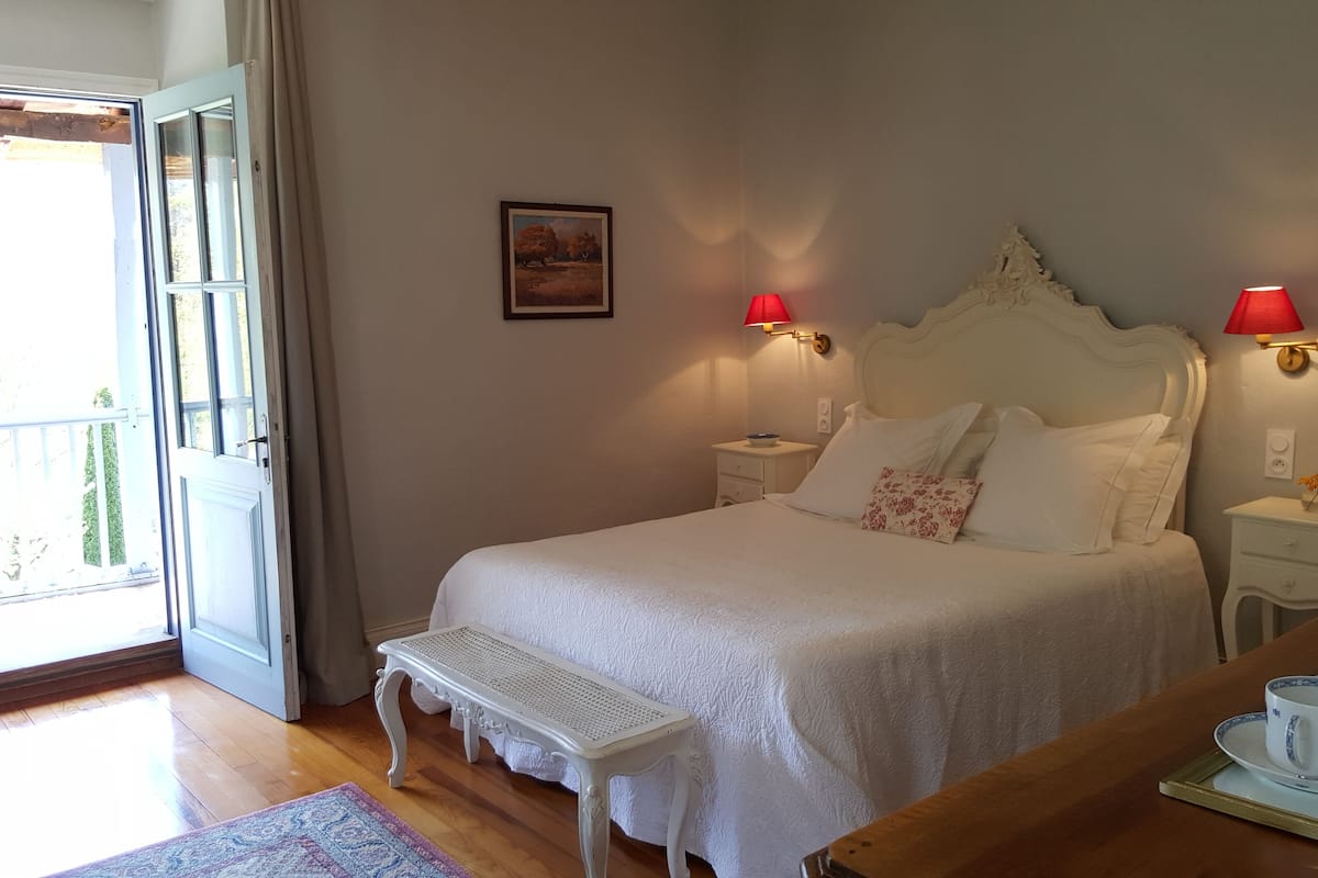 A bedroom with a large bed, a bench, a bedside table with a lamp, and a view of the outdoors.