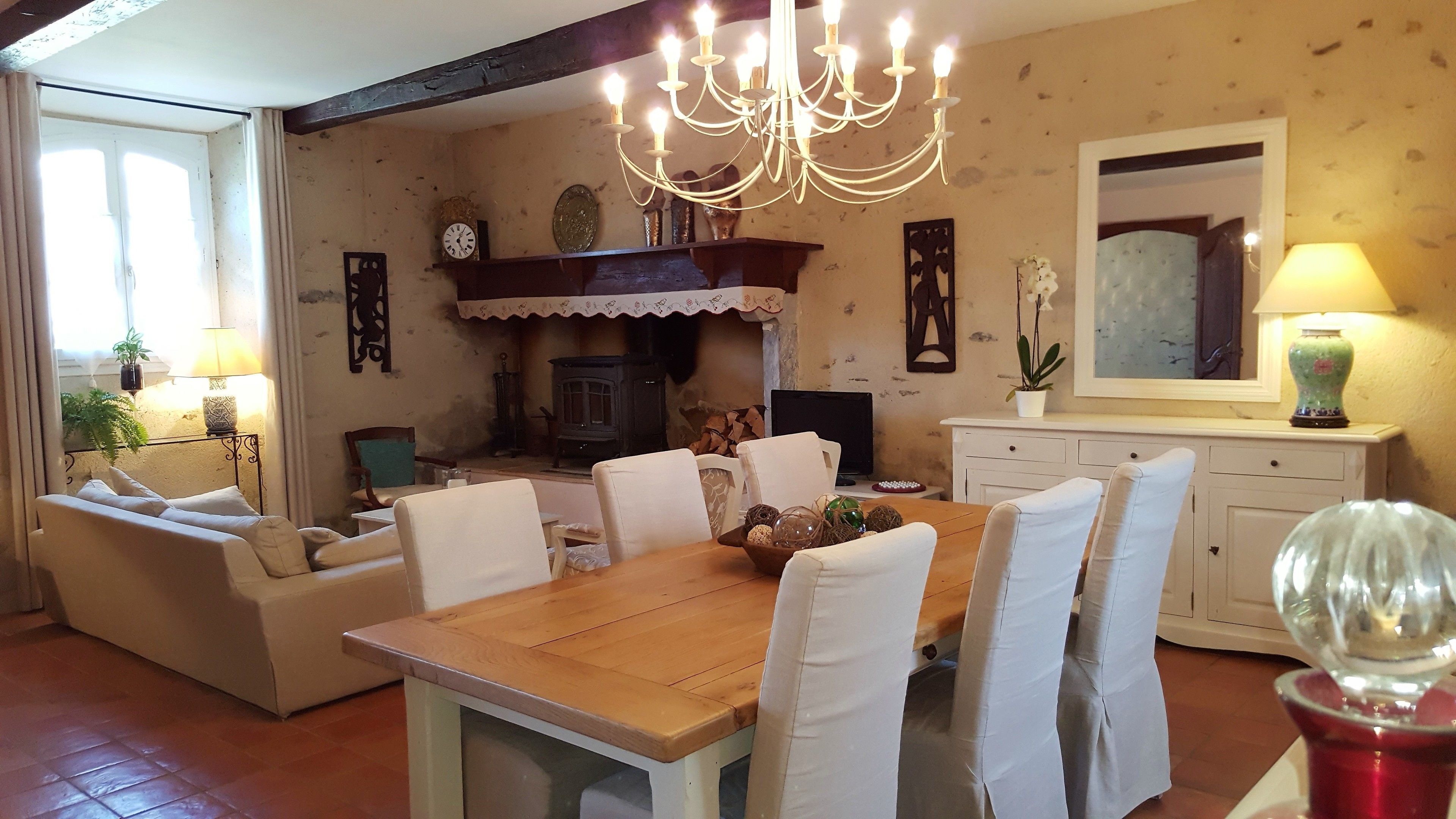 Une salle à manger avec une table en bois, des chaises blanches, un lustre et une cheminée.