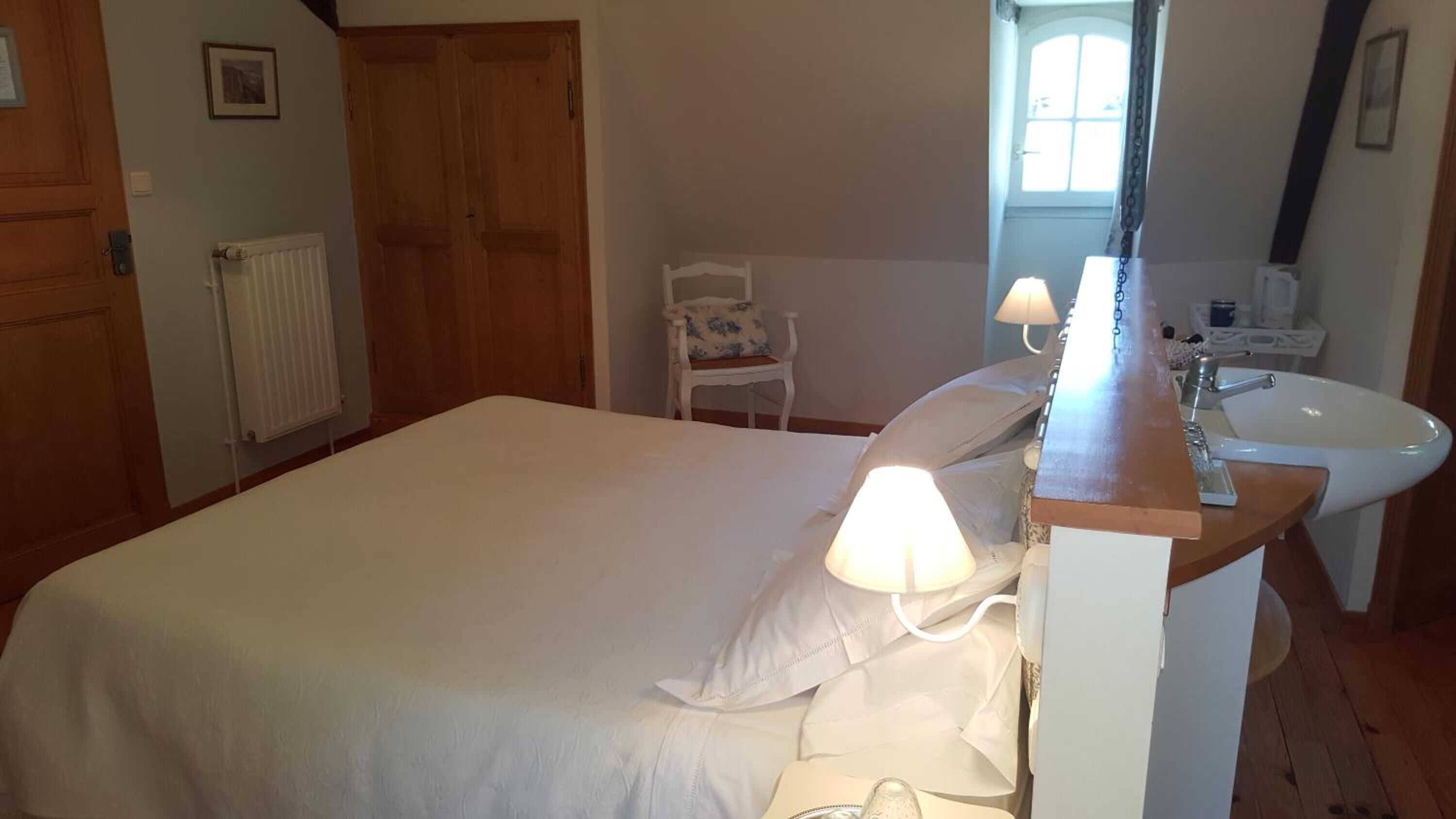 A bedroom with a bed, a chair, a bedside table with a lamp, and a bathroom with a sink and mirror.