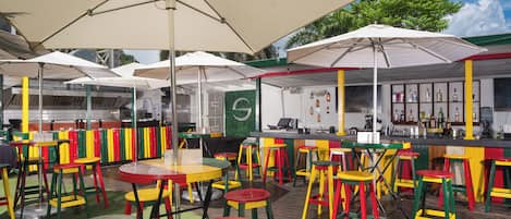 An outdoor dining area with colorful tables and chairs, umbrellas, and a bar area with a green wall and various bottles.