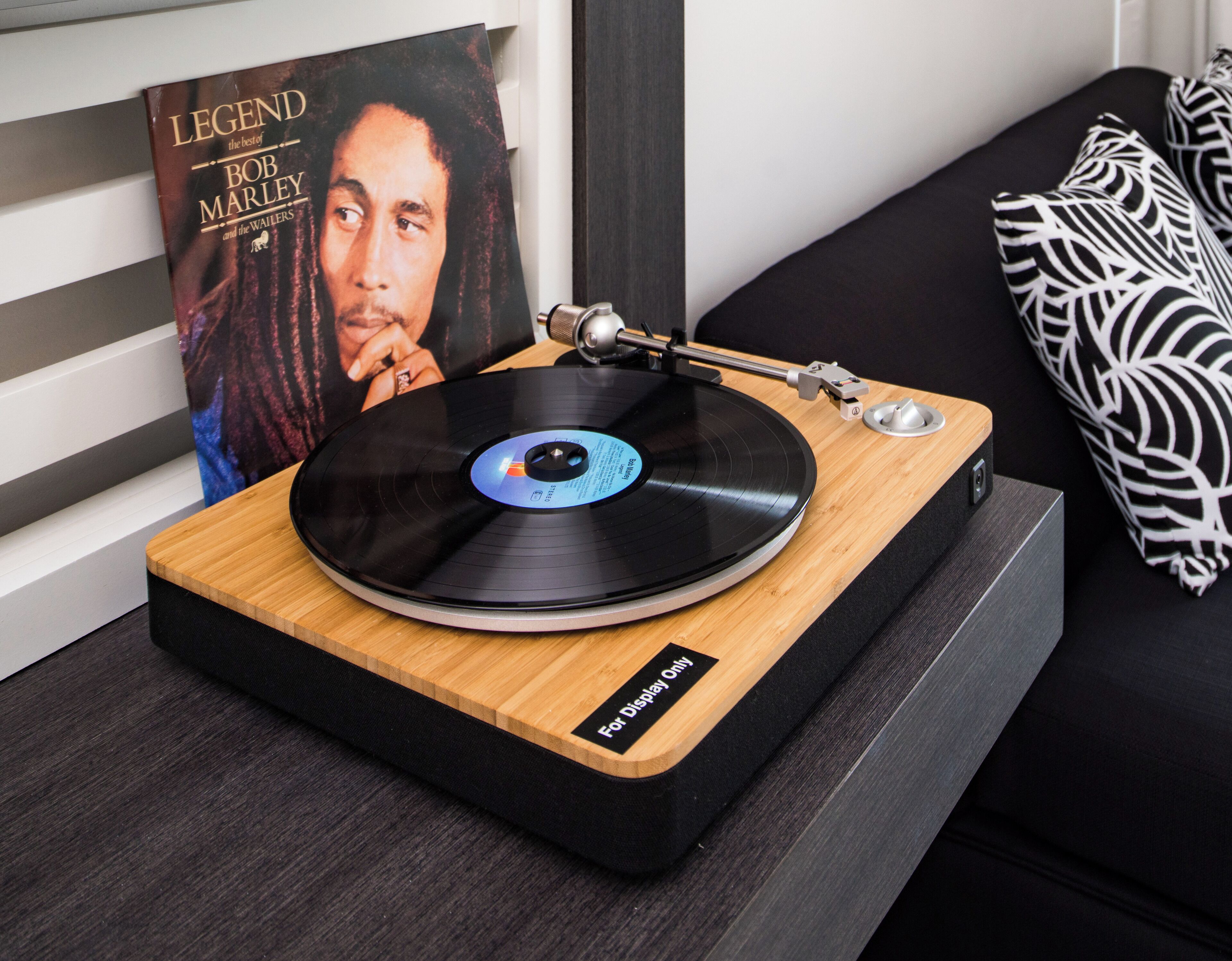 A turntable on a wooden platform with a record on it, a black and white patterned pillow, and a framed picture of Bob Marley on the wall.