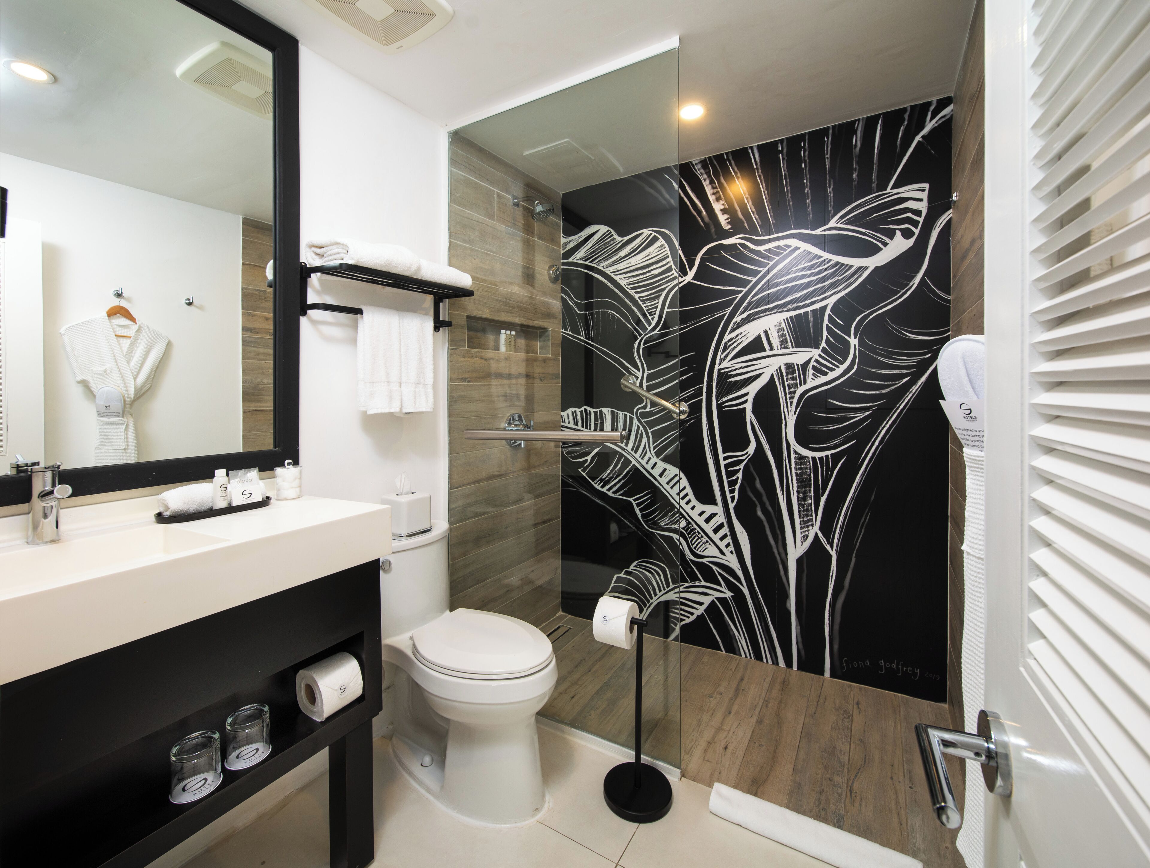 Un baño moderno con un espejo grande, un inodoro blanco, cabina de ducha con paredes de vidrio y un papel tapiz con estampado de hojas en blanco y negro.