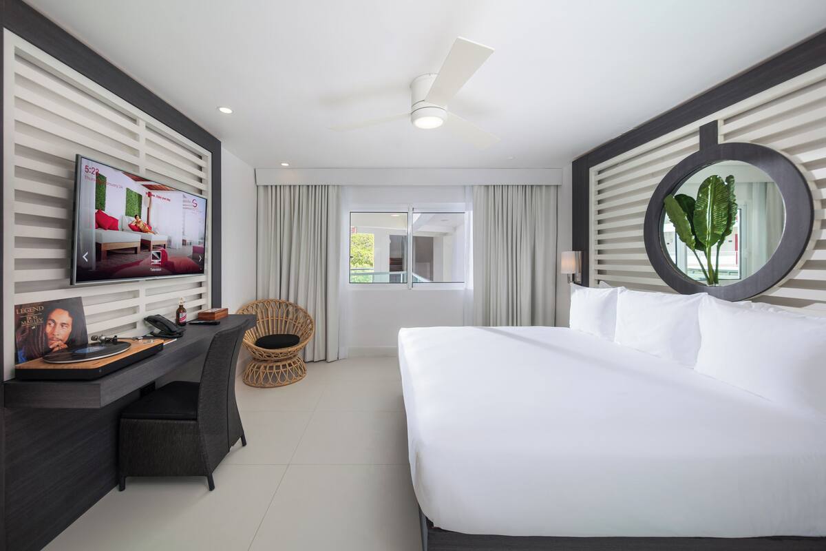 Una habitación de hotel moderna con una cama, un escritorio con una computadora y un televisor, una silla y un ventanal grande.