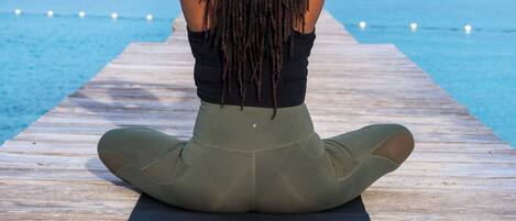 A person sitting on a yoga mat on a wooden pier, facing the sea.