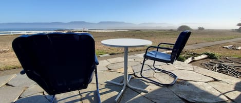 Two blue chairs and a white table on a stone patio with a view of the ocean and mountains.